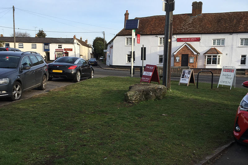 view of  cars parked by Fort End Green with House of Spice and The Cottage Bakery in the background 